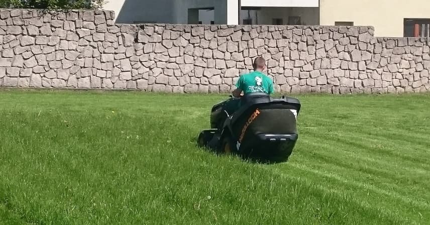 Mężczyzna kosi trawę traktorkiem na tle kamiennego muru i budynku.