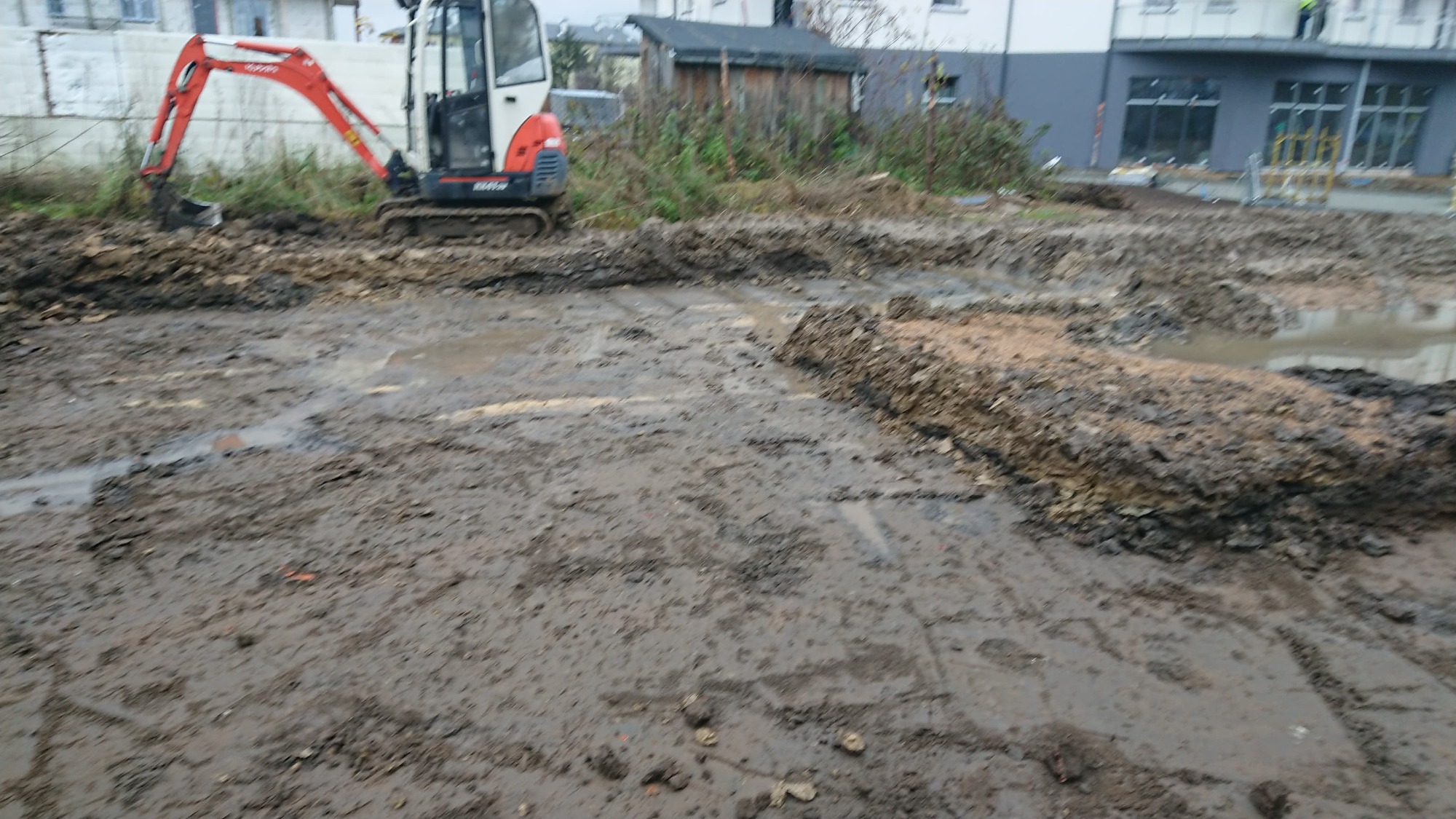 Mała koparka gąsienicowa na placu budowy, teren rozkopany i błotnisty, w tle budynek w trakcie budowy.