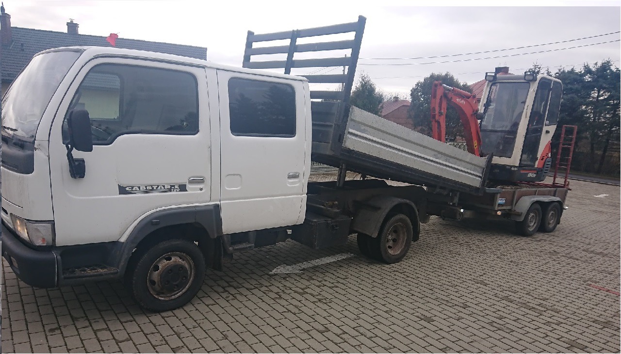 Biała wywrotka Nissan Cabstar-E z podniesioną skrzynią ładunkową, na której znajduje się pomarańczowa minikoparka na przyczepie, zaparkowana na brukowanej powierzchni.