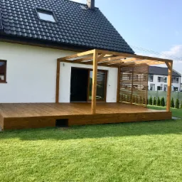 Nowoczesny dom z drewnianym tarasem i pergolą z lamelami, widok z zewnątrz na zadbany trawnik i inne budynki w tle.