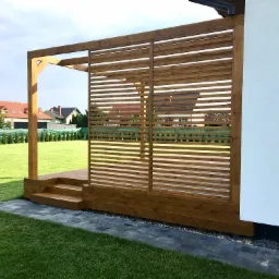 Drewniana konstrukcja z ażurową ścianą, tworząca zacienione przejście z trzema stopniami, przylegająca do białej ściany budynku, na tle zielonego trawnika i zabudowy jednorodzinnej.