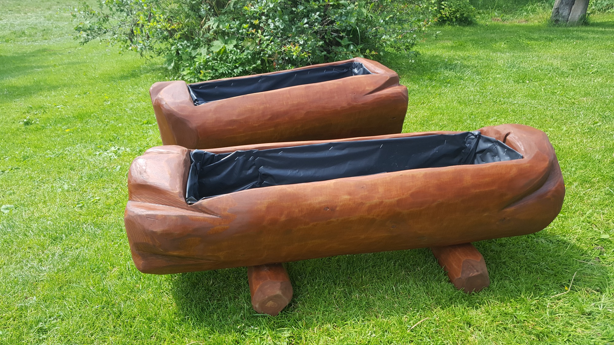 Dwa ręcznie rzeźbione, drewniane koryta na nóżkach, wyłożone czarną folią, stojące na trawniku w słoneczny dzień.