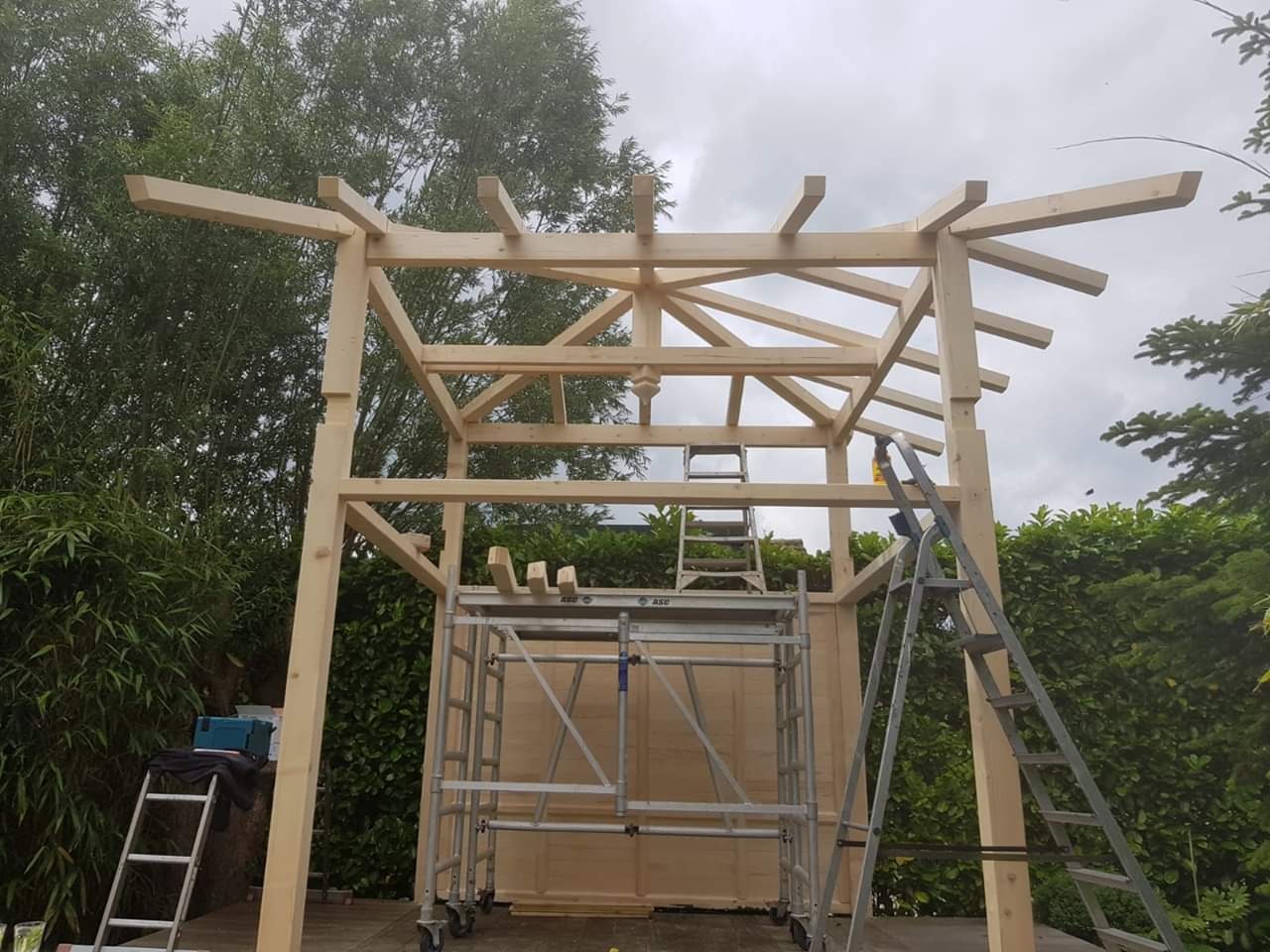 Surowa, drewniana konstrukcja altany w trakcie budowy, widoczne rusztowanie i drabiny, tło z zieleni ogrodowej i pochmurnego nieba.