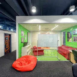 Nowoczesne biuro z ciemną wykładziną dywanową, czerwonym pufem i przeszklonym pokojem z zielonymi ścianami, widoczne oświetlenie sufitowe i designerskie drzwi.