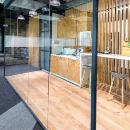 Jasna drewniana podłoga w biurze, widok przez szklaną ścianę na aneks kuchenny z drewnianymi szafkami i wysokim stołem barowym z drewnianymi krzesłami, widoczny fragment wykładziny dywanowej.