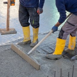 FULER Sp. z o.o. - Dwóch robotników w żółtych gumowcach rozprowadza mokry beton na zbrojeniu fundamentów za pomocą łaty i poziomicy w trakcie wylewania posadzki.