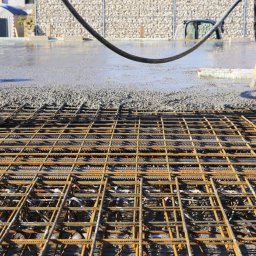 FULER Sp. z o.o. - Zbrojenie fundamentu z siatki stalowej przed wylaniem betonu, widoczne gumowe buty robotników w tle i wąż do betonu, ściana z kamieni w koszach gabionowych w tle.