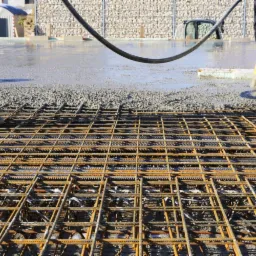 Zbrojenie fundamentu z siatki stalowej przed wylaniem betonu, widoczne gumowe buty robotników w tle i wąż do betonu, ściana z kamieni w koszach gabionowych w tle.