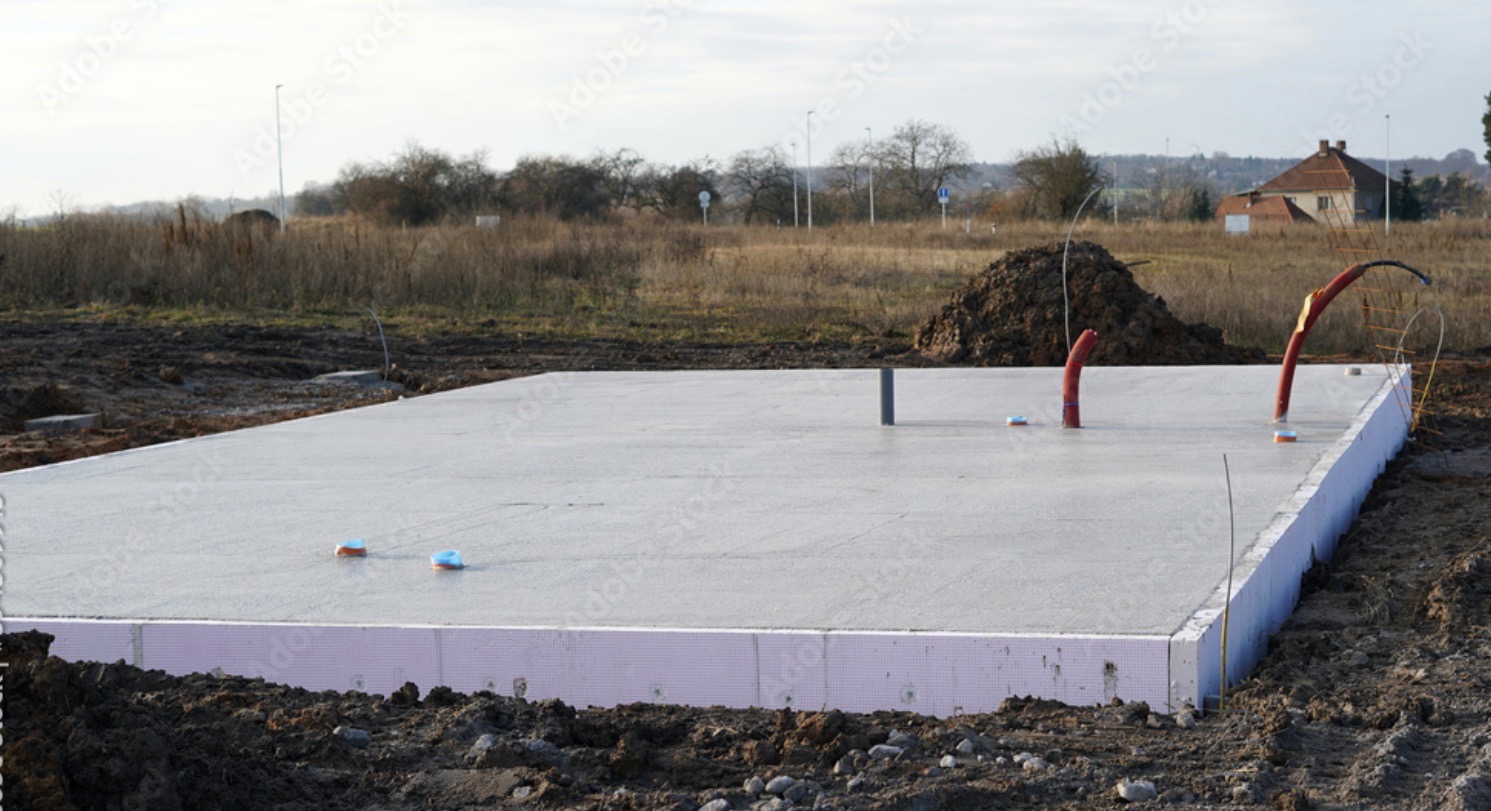 Wylana betonowa płyta fundamentowa z izolacją pionową, wystające rury instalacyjne, widok na pole i zabudowania w tle.