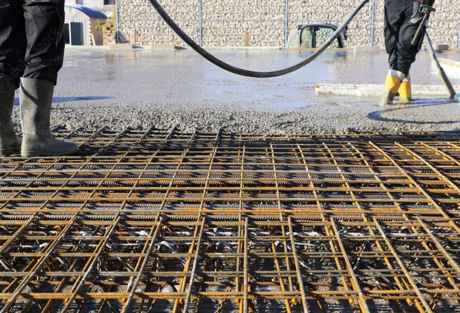 Zbrojenie fundamentu z siatki stalowej przed wylaniem betonu, widoczne gumowe buty robotników w tle i wąż do betonu, ściana z kamieni w koszach gabionowych w tle.