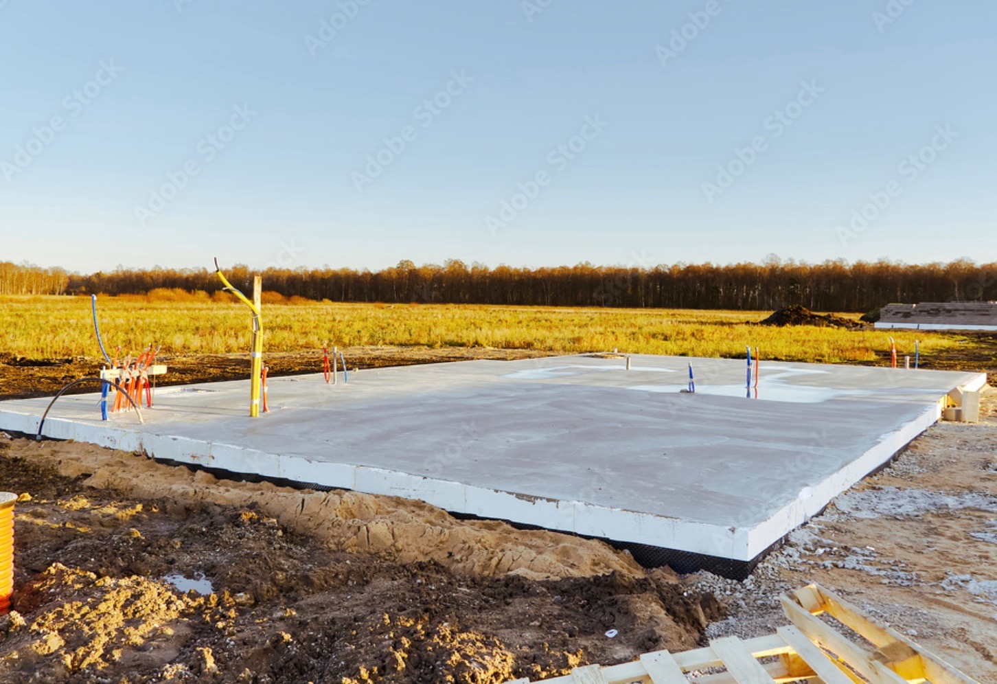 Płyta fundamentowa z instalacjami pionowymi, ocieplona styropianem, na tle krajobrazu z polami i lasem.