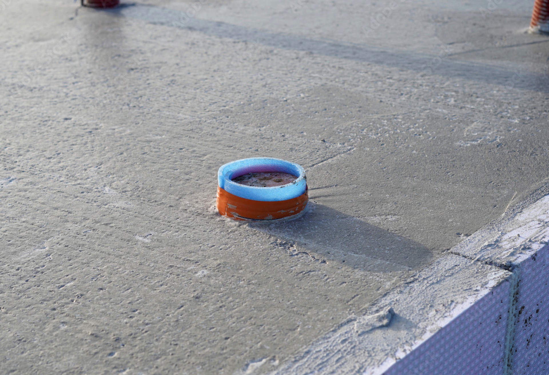 Świeżo wylany beton fundamentowy z wystającą rurą instalacyjną w pomarańczowo-niebieskiej osłonie ochronnej i widoczną izolacją obwodową.