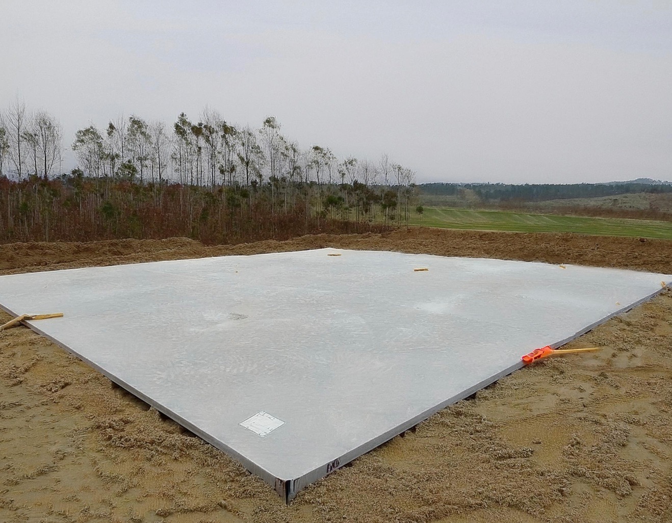 Wylana, gładka płyta fundamentowa na piaszczystym podłożu z widocznymi żółtymi znacznikami i pomarańczowym narzędziem, w tle drzewa i pole pod pochmurnym niebem.