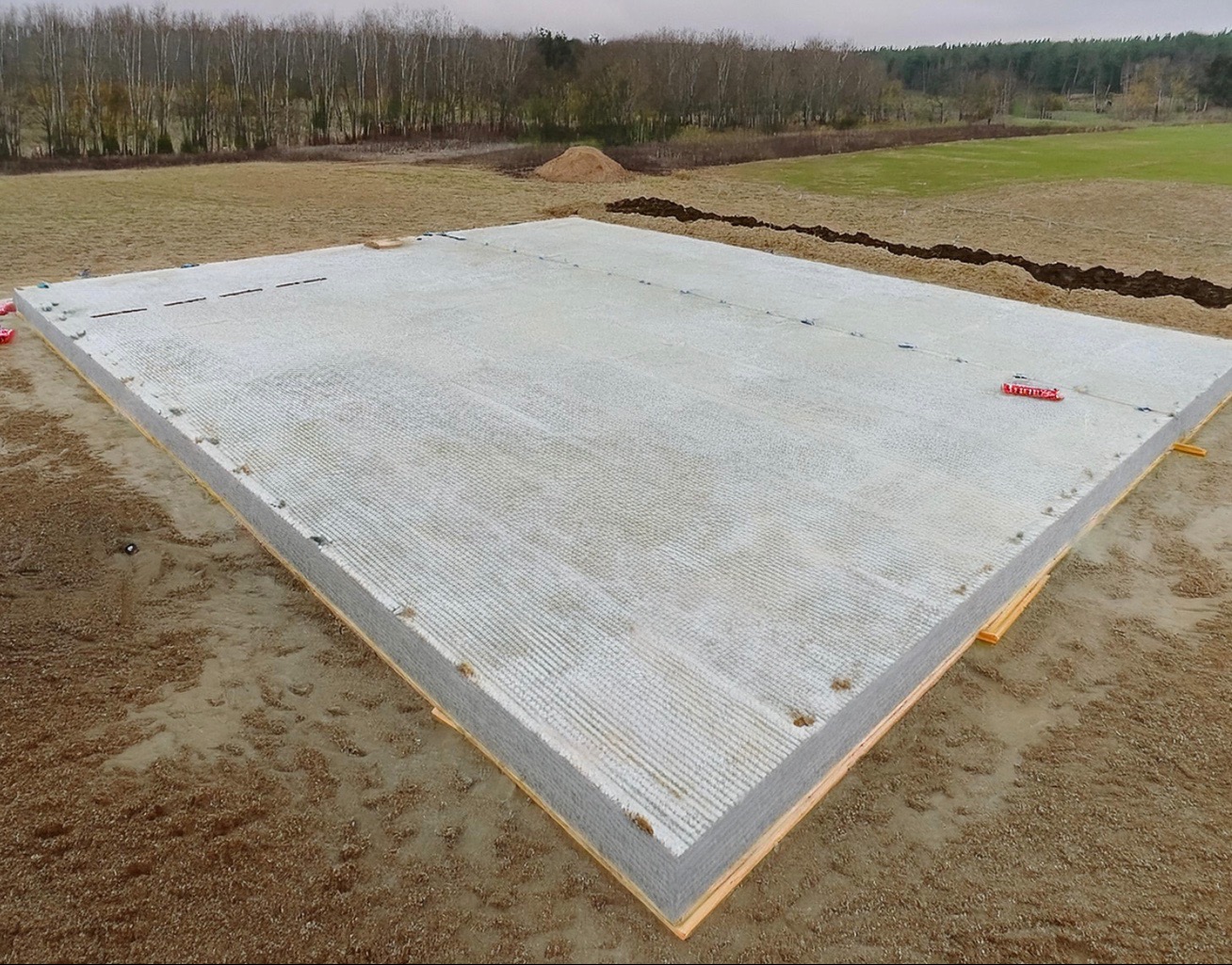 Przygotowany, betonowy fundament płyty pod budynek, z widoczną izolacją pionową, na tle pola i lasu w pochmurny dzień.