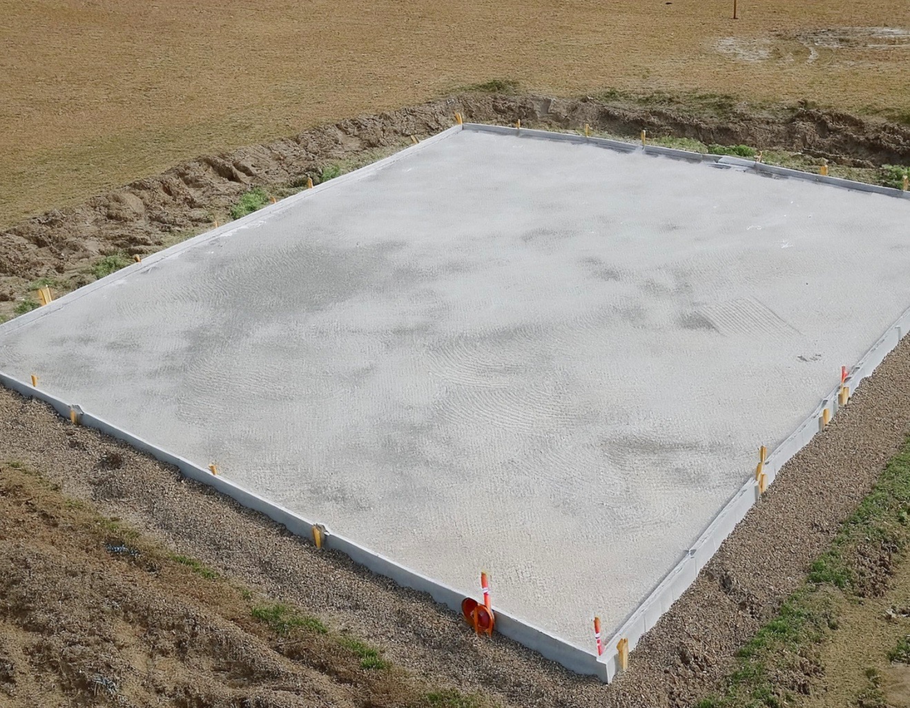 Wylana, świeża betonowa płyta fundamentowa z widocznymi żółtymi znacznikami poziomów i pomarańczowym elementem zabezpieczającym w rogu, otoczona ziemią i żwirem.