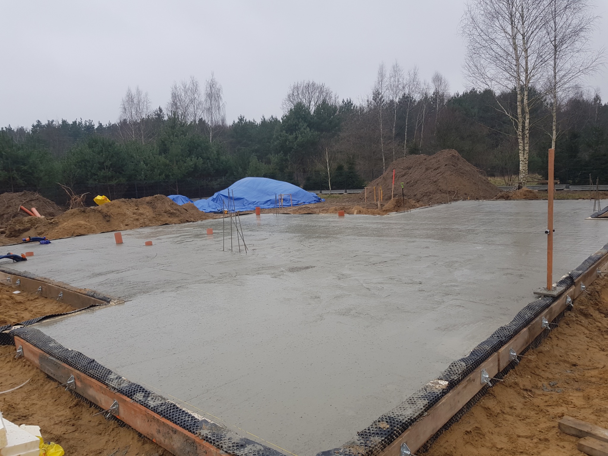 Wylany betonowy fundament płyty, widoczne szalunki z membraną kubełkową, w tle hałda ziemi przykryta plandeką i zalesiony teren.
