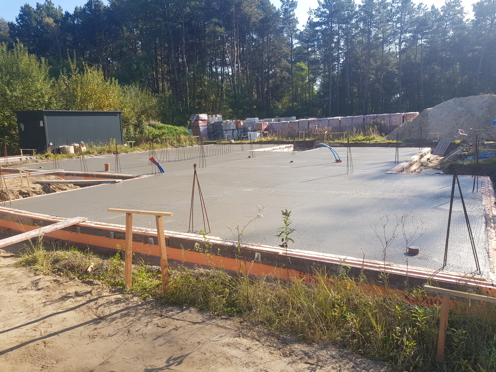 Wylany fundament pod budynek z widocznym szalunkiem, zbrojeniem i stertą cegieł w tle, w słoneczny dzień.