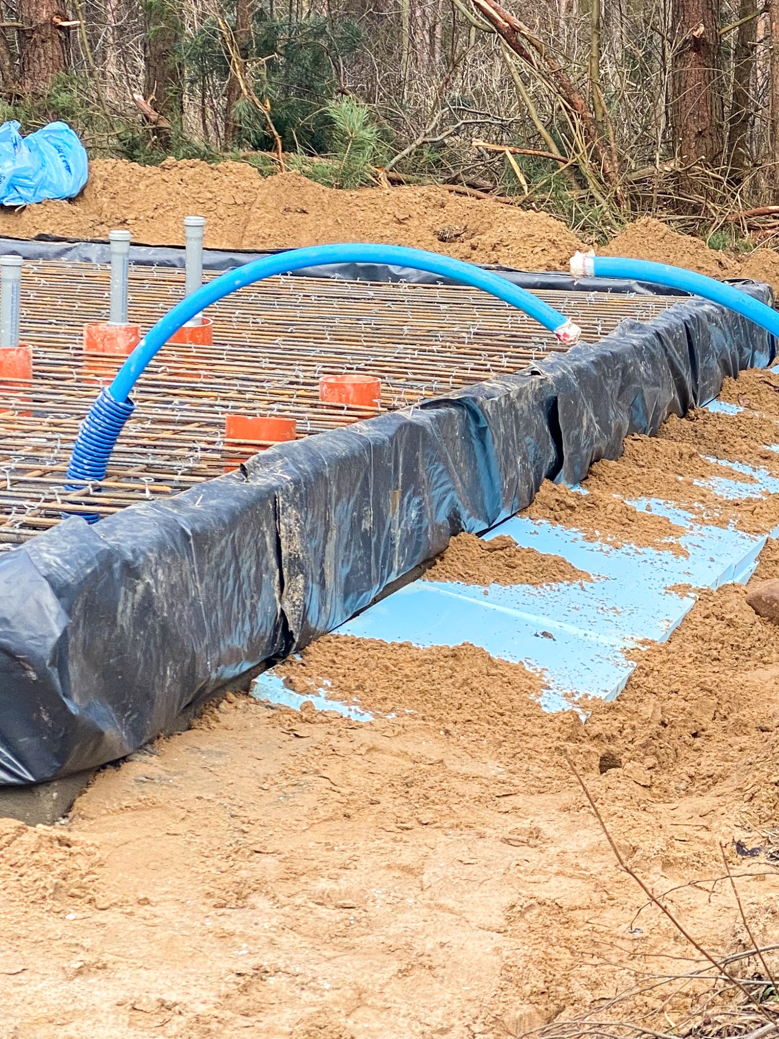 Przygotowanie zbrojenia fundamentów z niebieską rurą peszel i styropianem izolacyjnym zabezpieczonym folią w wykopie, widok na las w tle.