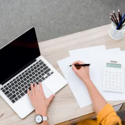 Widok z góry na biurko z laptopem, kalkulatorem, telefonem i ręką piszącą na papierze, sugerujący pracę biurową.