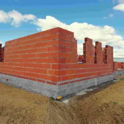 Wznoszenie ścian parteru z czerwonej cegły na fundamencie z betonowych bloczków, widoczne otwory w cegłach i żółta rura kanalizacyjna wystająca z ziemi, w tle zielone pole i błękitne niebo...