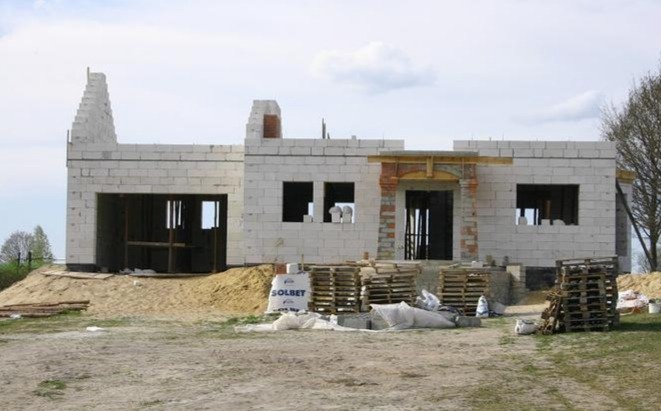 Niedokończony budynek z białych bloczków, widoczne palety i worki z zaprawą, wstępny etap budowy z niezadaszonymi otworami okiennymi i drzwiowymi.