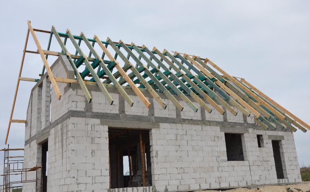 Surowy budynek z białych pustaków z drewnianą konstrukcją dachu w trakcie montażu, widoczne zielone elementy impregnatu na belkach, szare niebo w tle.