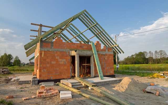 Surowy stan domu jednorodzinnego z czerwonej cegły, z częściowo wykonaną konstrukcją dachu z zielonych belek, widoczne bloczki i deski na ziemi.
