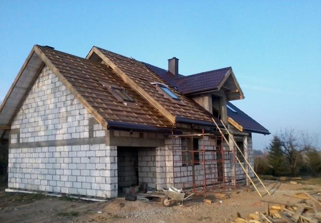 Dom w trakcie budowy z białych bloczków, częściowo pokryty brązowym dachem z oknami dachowymi, widoczne rusztowanie i materiały budowlane na placu.