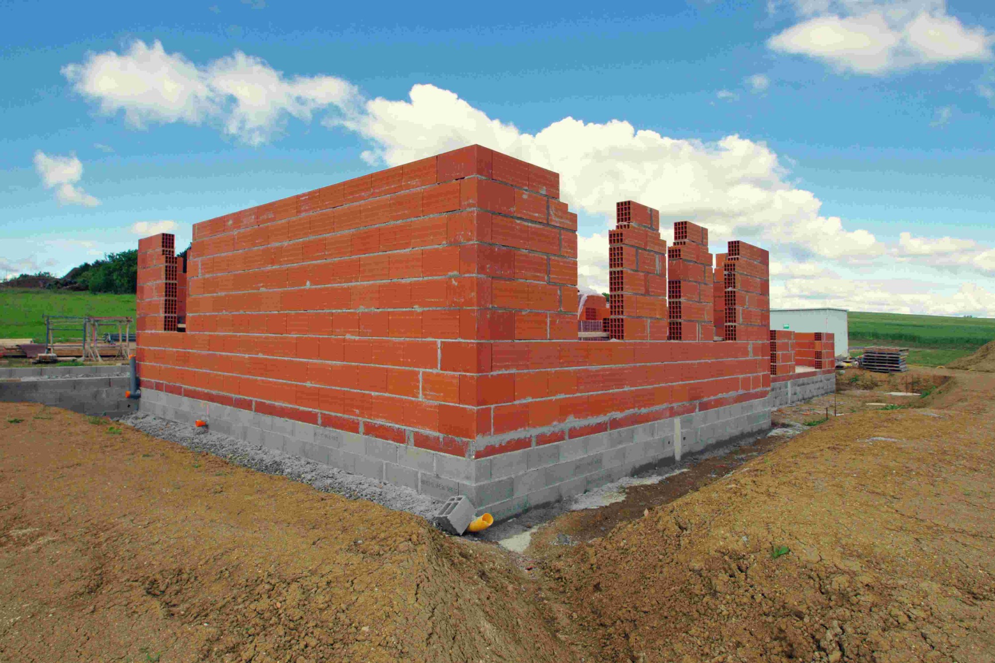Wznoszenie ścian parteru z czerwonej cegły na fundamencie z betonowych bloczków, widoczne otwory w cegłach i żółta rura kanalizacyjna wystająca z ziemi, w tle zielone pole i błękitne niebo...