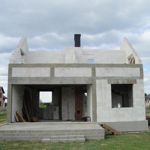 Surowy stan domu z białych bloczków, widoczne otwory okienne i drzwiowe, komin z czarnym wkładem, podmurówka z betonu, składowane deski i bloczki wewnątrz.