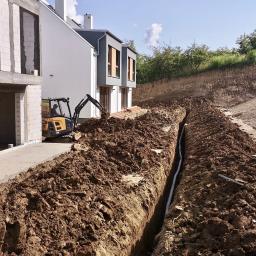 Zaprojektuj i Wybuduj Sp. z o.o. - Koparka Volvo EC15E przy wykopie pod rurę kanalizacyjną na tle budynków w stanie surowym otwartym.