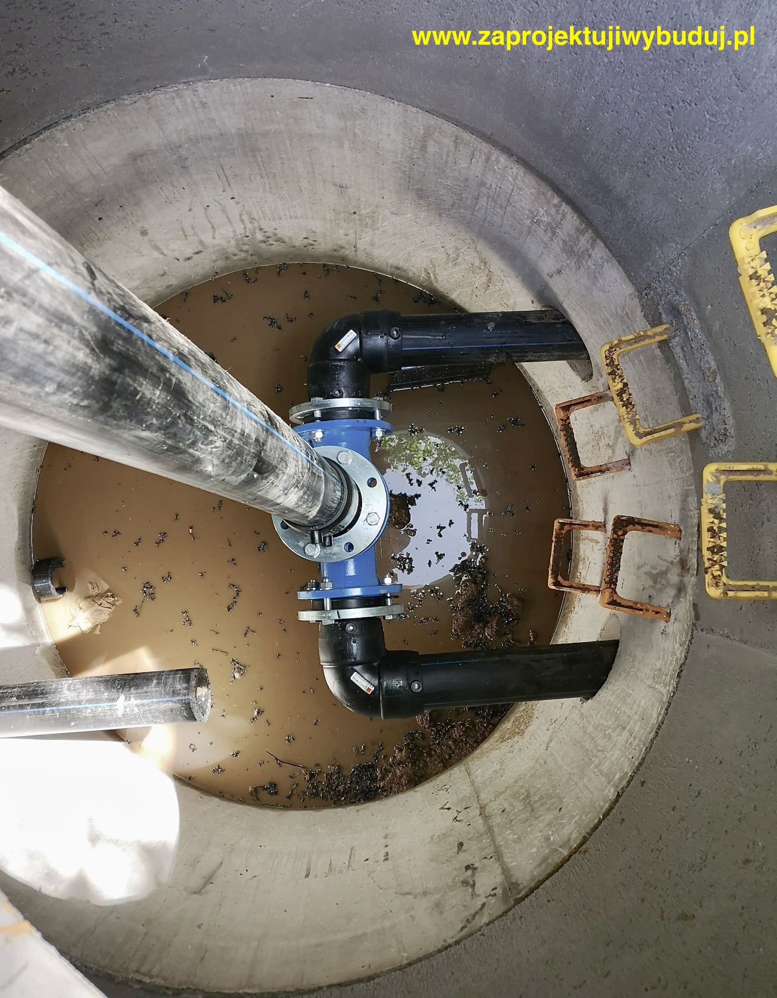 Wnętrze betonowej studni rewizyjnej z zainstalowanym trójnikiem i rurami z tworzywa sztucznego, częściowo zanurzonymi w brudnej wodzie. Widoczne żółte stopnie drabiny.