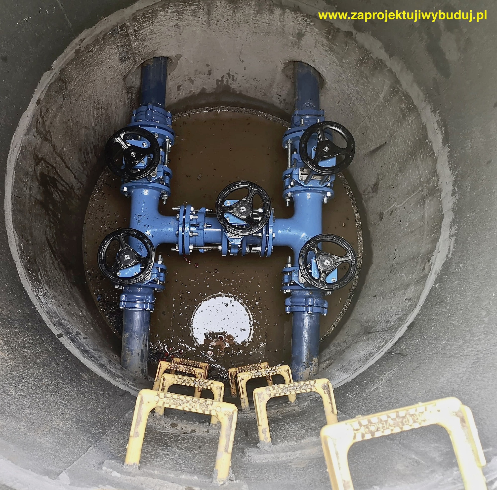 Wnętrze betonowej studni z niebieską instalacją wodociągową z zaworami, częściowo zanurzoną w wodzie, z żółtymi stopniami drabinkowymi na dnie.