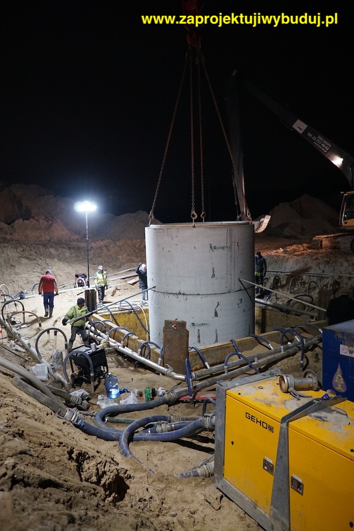 Montaż betonowych kręgów studni przy użyciu dźwigu nocą, widoczne pompy odwadniające i pracownicy w odblaskowych kamizelkach.