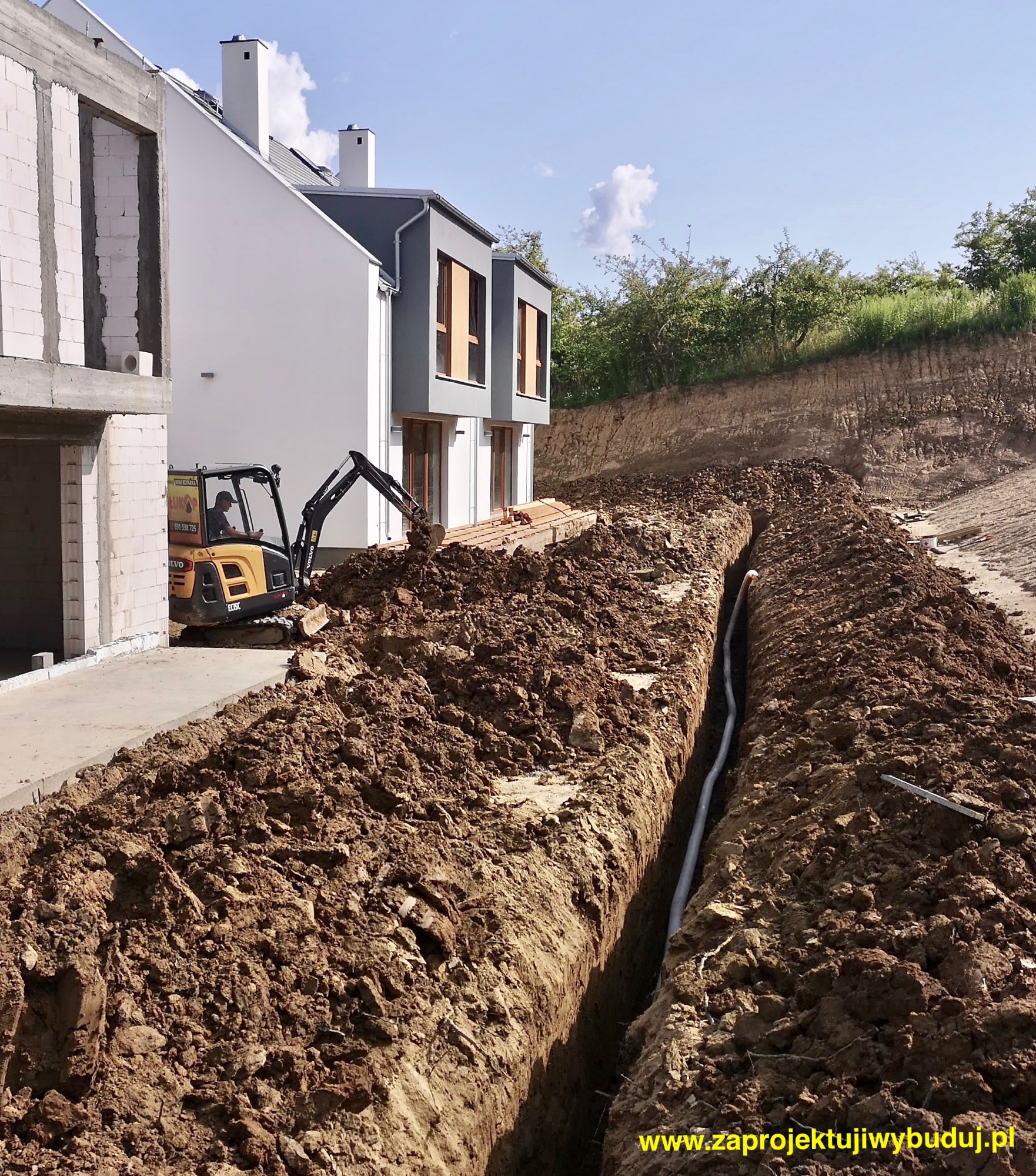 Koparka Volvo EC15E przy wykopie pod rurę kanalizacyjną na tle budynków w stanie surowym otwartym.