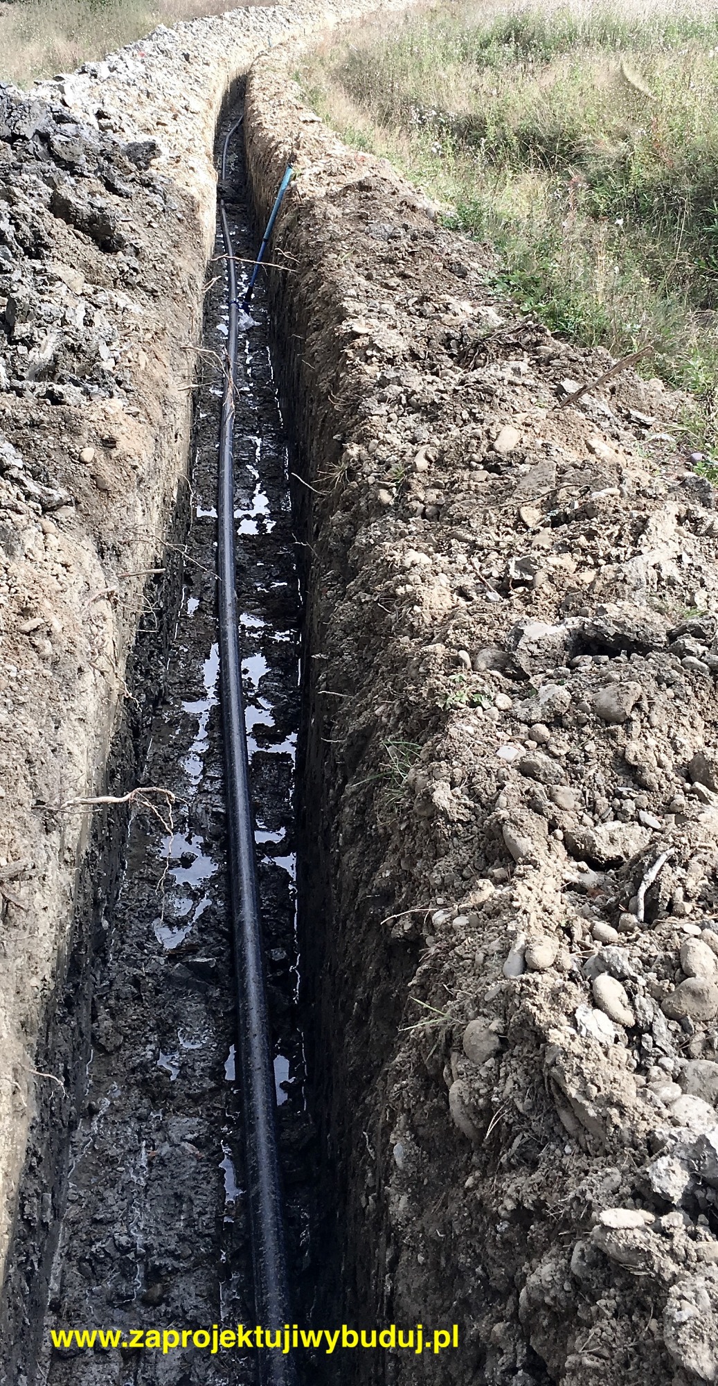 Wykop z ułożoną czarną rurą instalacji sanitarnej, częściowo wypełniony wodą, widok z góry.