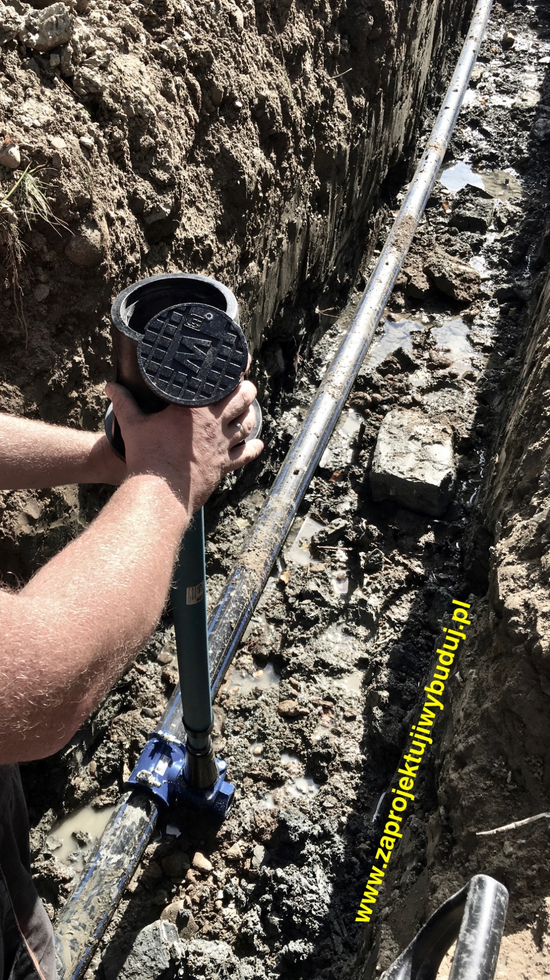 Montaż studzienki wodomierzowej w wykopie, widoczna rura wodociągowa i narzędzia, teren budowy.