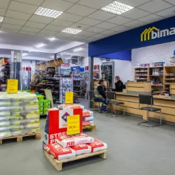 Wnętrze składu budowlanego w Rzeszowie z widocznym regałem z materiałami budowlanymi, paletami z workami zaprawy i kasą z klientami.