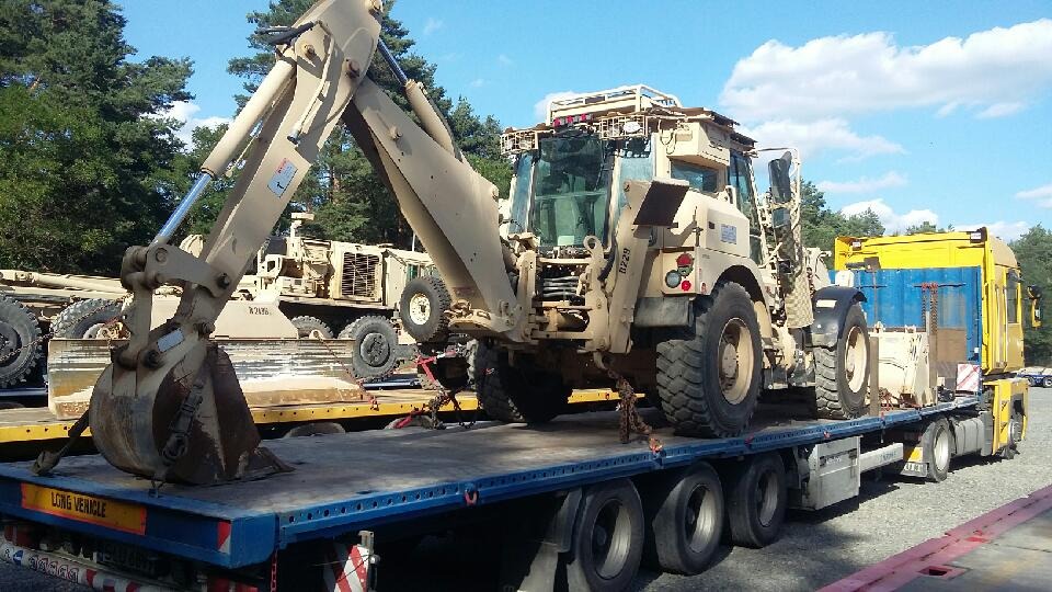 Beżowa koparko-ładowarka wojskowa zabezpieczona łańcuchami na niskopodwoziowej naczepie ciężarówki, w tle widoczna kolejna maszyna budowlana i drzewa.