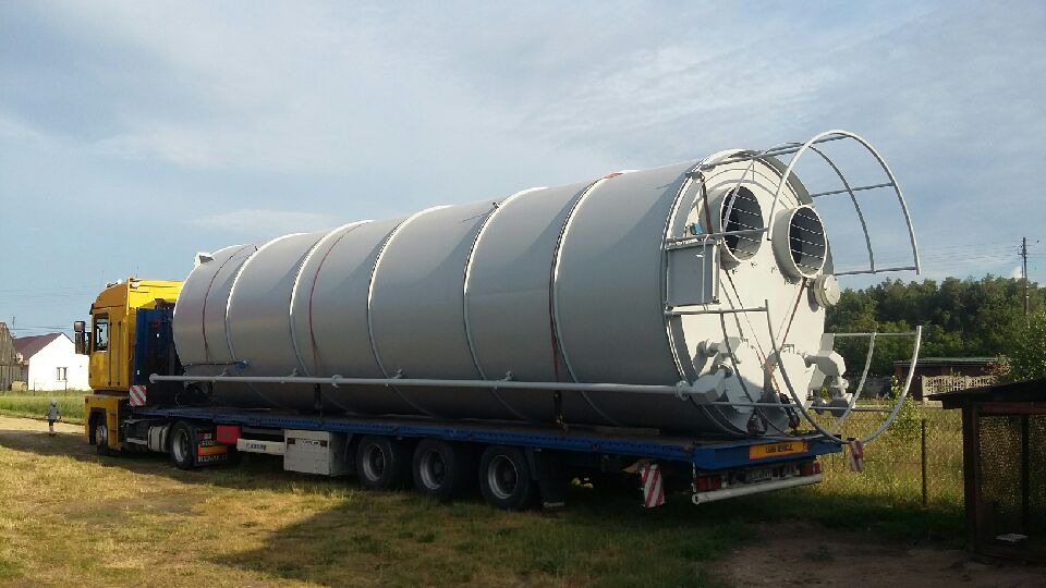 Żółta ciężarówka Renault Magnum przewozi na niskopodwoziowej naczepie duży, szary, cylindryczny zbiornik z metalową konstrukcją na jednym końcu.