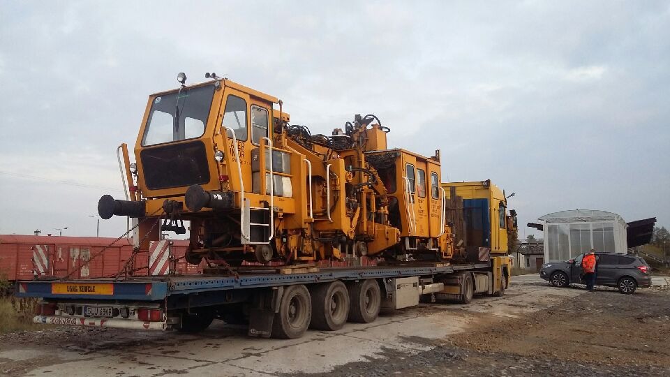 Żółta maszyna torowa zabezpieczona łańcuchami na niskopodwoziowej naczepie ciężarówki, widoczny wagon kolejowy w tle.