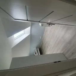 Wnętrze poddasza po remoncie, z widocznym oknem dachowym, świeżo położoną podłogą z paneli imitujących drewno i pomalowanymi na szaro-biało ścianami, w tym skośnymi, oraz minimalistyczną szafką...