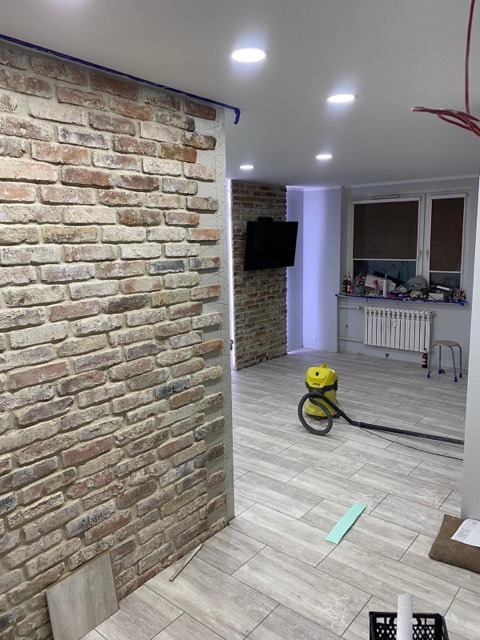 Ściana z cegły dekoracyjnej w trakcie montażu, widoczne zabezpieczenia taśmą malarską i poziomowanie, podłoga wyłożona płytkami imitującymi drewno, odkurzacz przemysłowy w tle.