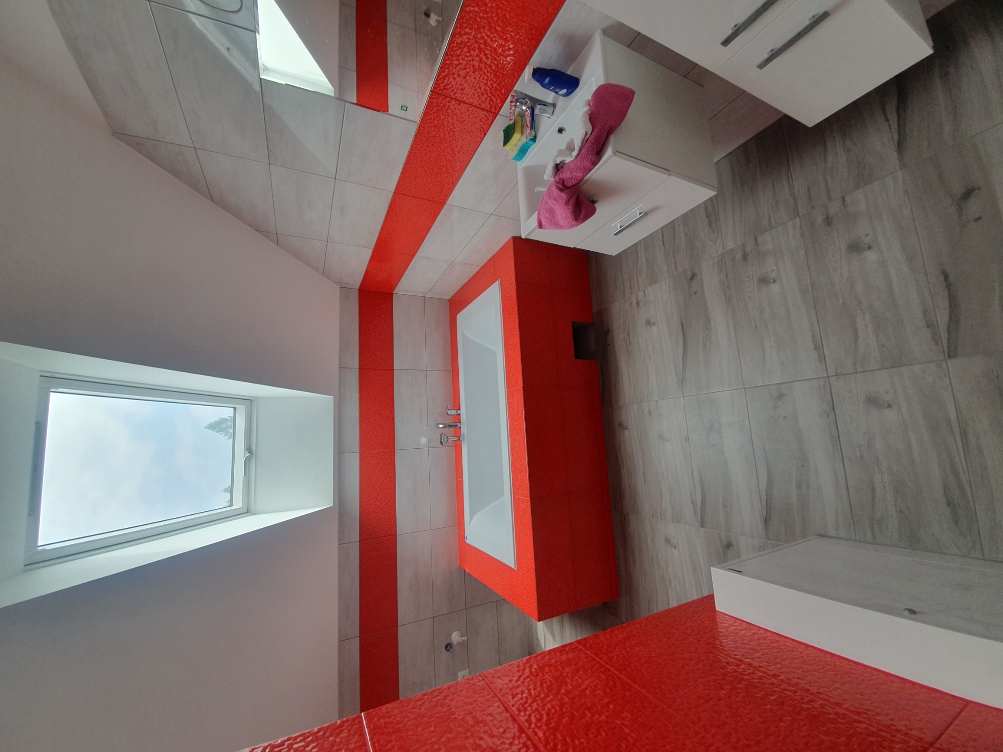 Wykończona łazienka z akcentami czerwonej mozaiki na ścianach i obudowie wanny, płytki podłogowe imitujące drewno, biała szafka pod umywalką i okno dachowe w skosie.