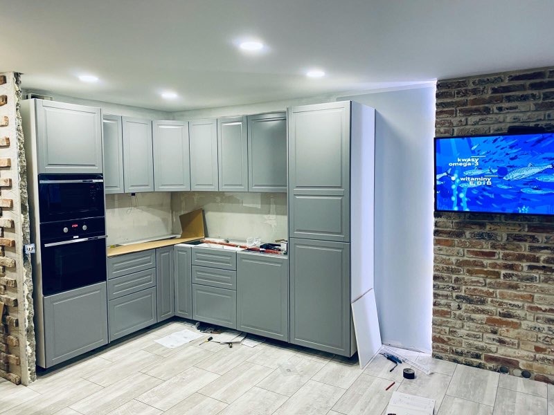 Montaż szarej zabudowy kuchennej z piekarnikiem w trakcie wykończenia, widoczny fragment ceglanej ściany i telewizor z motywem ryb