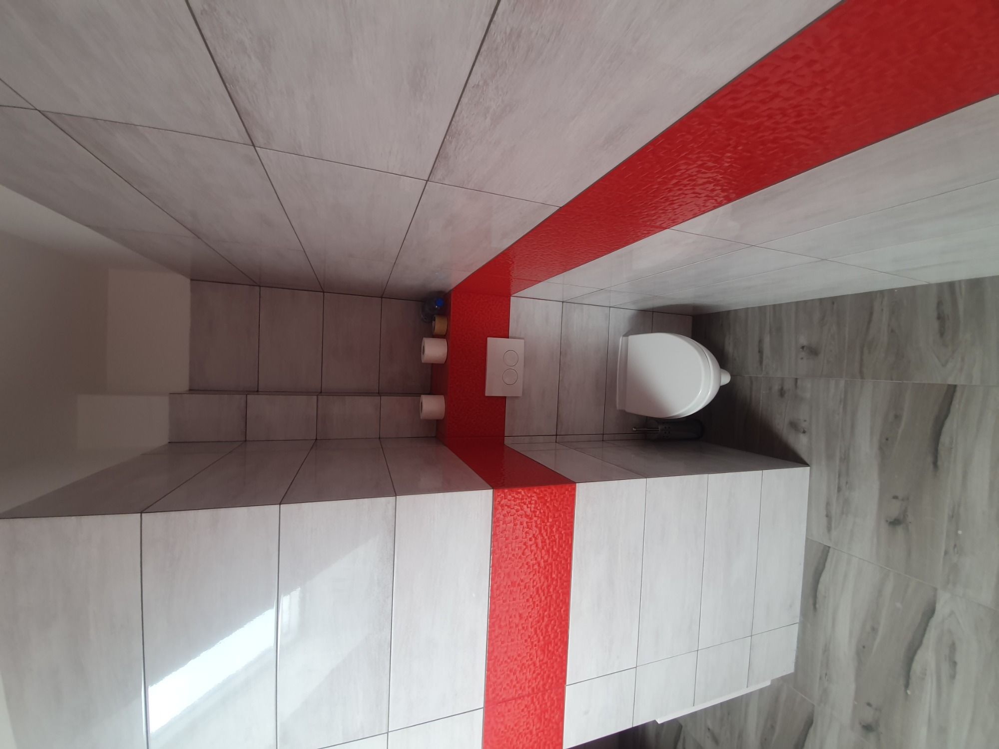 Nowoczesna łazienka z jasnymi płytkami i szerokim, czerwonym pasem dekoracyjnym, obejmującym podłogę i ścianę, widoczna toaleta podwieszana i zapas papieru toaletowego.