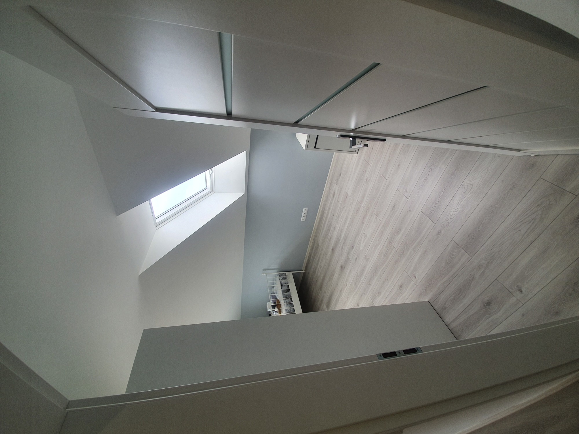 Wnętrze poddasza po remoncie, z widocznym oknem dachowym, świeżo położoną podłogą z paneli imitujących drewno i pomalowanymi na szaro-biało ścianami, w tym skośnymi, oraz minimalistyczną szafką...