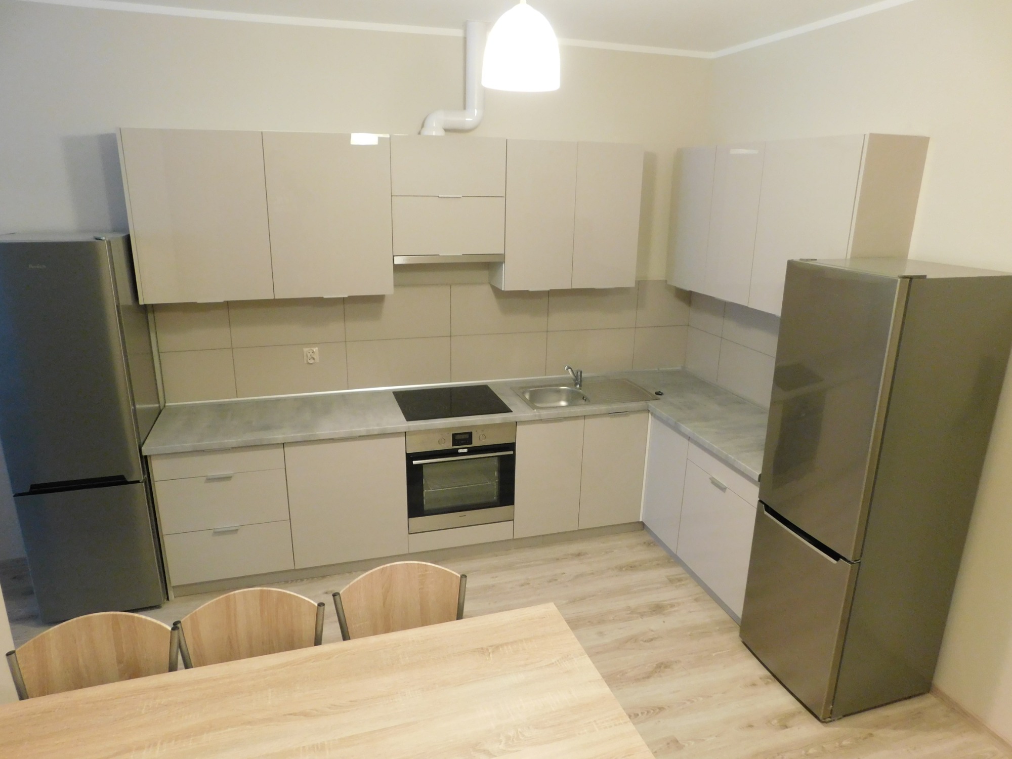 Jasna, nowoczesna kuchnia z beżowymi szafkami, szarą lodówką i drewnianym stołem, oświetlona pojedynczą lampą wiszącą.