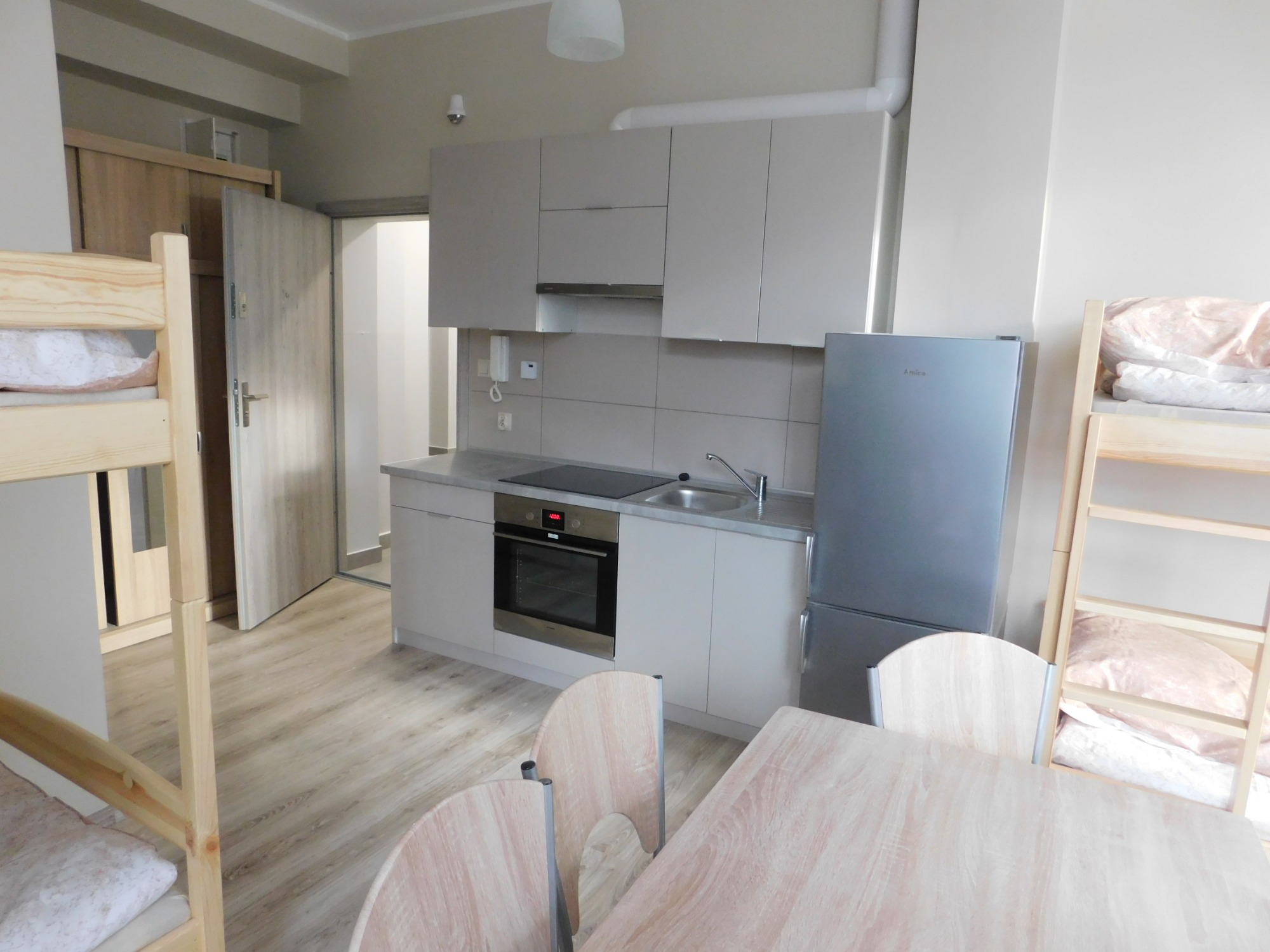 Wnętrze pokoju z aneksem kuchennym: drewniane łóżko piętrowe, stół z krzesłami oraz zabudowa kuchenna z płytą indukcyjną i lodówką.