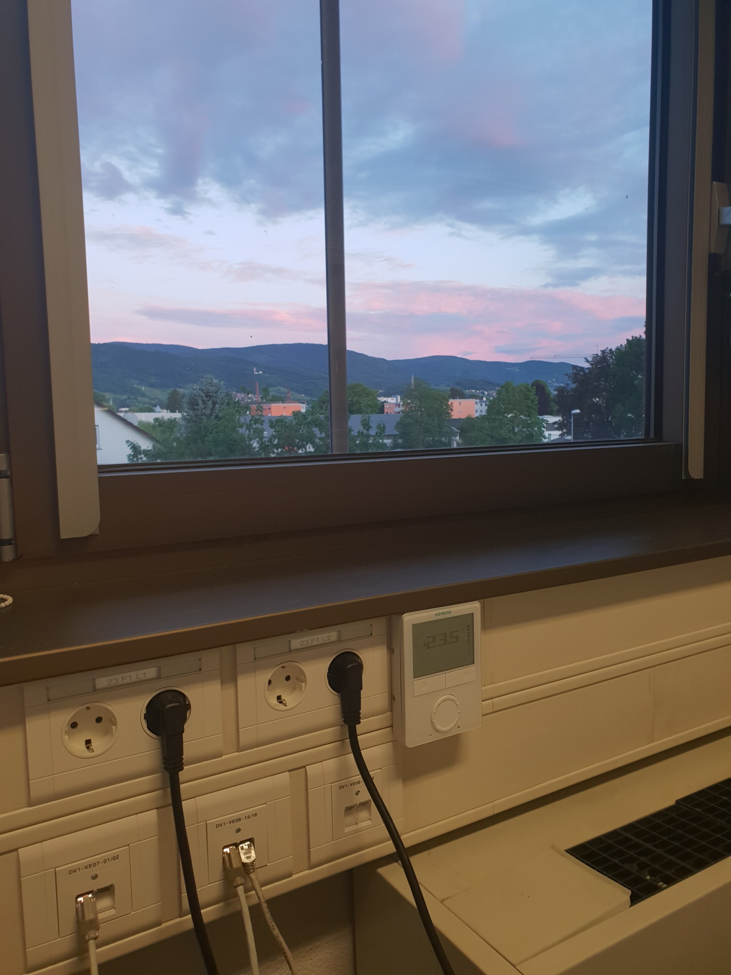 Widok z okna na miasto i góry o zmierzchu, z widocznymi gniazdkami elektrycznymi i sieciowymi oraz termostatem Siemens na ścianie poniżej.